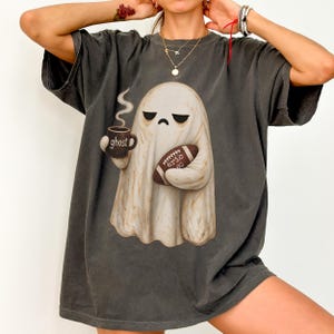 May include: A charcoal gray short-sleeve t-shirt featuring a graphic of a beige ghost with a grumpy expression. The ghost holds a dark mug labeled ghost and a brown football with the text eric 80. The shirt has a crew neckline and loose fit.