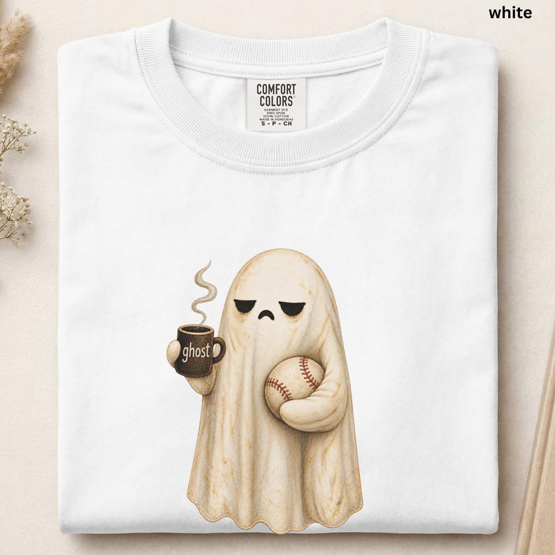 May include: A white crew neck t-shirt featuring a graphic of a beige ghost with a sad expression. The ghost holds a brown mug with the word ghost printed on it and a baseball in its other hand. The shirt has a Comfort Colors brand label inside.