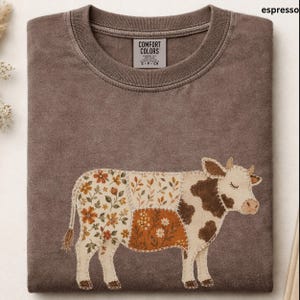 May include: An espresso-colored crew neck t-shirt featuring a cow graphic composed of patchwork-style fabric segments. The cow design includes sections with floral patterns in orange, brown, and cream tones, and a label reading Comfort Colors.