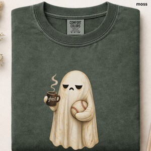 May include: A moss green crewneck t-shirt featuring a graphic of a cream-colored ghost with a sad expression. The ghost holds a brown mug labeled ghost with steam rising and a white baseball with red stitching in its other hand.