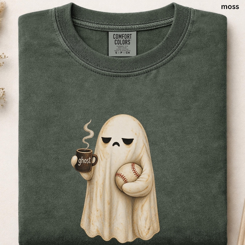 May include: A moss green crewneck t-shirt featuring a graphic of a cream-colored ghost with a sad expression. The ghost holds a brown mug labeled ghost with steam rising and a white baseball with red stitching in its other hand.