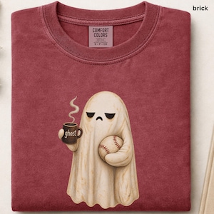 May include: A brick red t-shirt featuring a graphic of a cream-colored ghost with a sad expression, holding a brown mug with the word ghost printed on it and a baseball. The shirt has a Comfort Colors brand label inside the collar area.