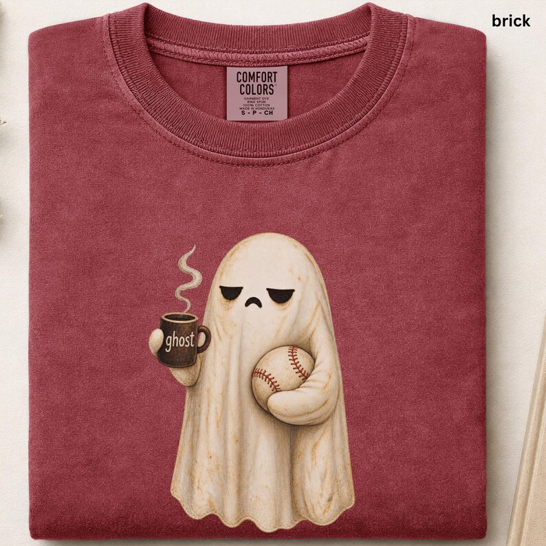 May include: A brick red t-shirt featuring a graphic of a cream-colored ghost with a sad expression, holding a brown mug with the word ghost printed on it and a baseball. The shirt has a Comfort Colors brand label inside the collar area.