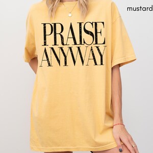 May include: Mustard-colored t-shirt with the words "PRAISE ANYWAY" in large, black, bold letters. The shirt has short sleeves and a relaxed fit. The model is wearing a gold necklace and a red bracelet.