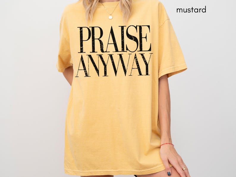 May include: Mustard-colored t-shirt with the words "PRAISE ANYWAY" in large, black, bold letters. The shirt has short sleeves and a relaxed fit. The model is wearing a gold necklace and a red bracelet.