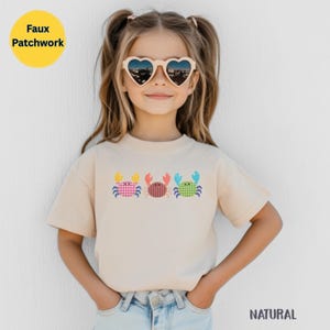 Puede incluir: Una camiseta de manga corta de color beige con un estampado gráfico de tres cangrejos en fila. Los cangrejos están diseñados con un patrón de imitación de patchwork en colores rosa, naranja y verde. La camiseta tiene cuello redondo.