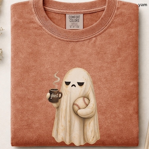 May include: A rust-colored crew neck t-shirt featuring a graphic of a sad-faced ghost holding a steaming mug labeled ghost and a baseball. The shirt has a Comfort Colors brand tag at the inner neckline and a textured, washed-out fabric appearance.