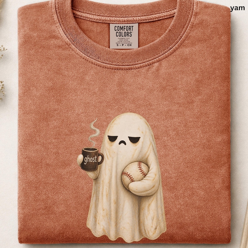 May include: A rust-colored crew neck t-shirt featuring a graphic of a sad-faced ghost holding a steaming mug labeled ghost and a baseball. The shirt has a Comfort Colors brand tag at the inner neckline and a textured, washed-out fabric appearance.