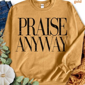 May include: Mustard-colored sweatshirt with the words "PRAISE ANYWAY" printed in large, black, block letters. The sweatshirt has a crew neck and long sleeves. The fabric appears soft and comfortable.