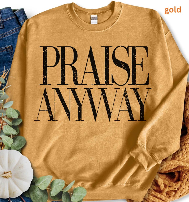 May include: Mustard-colored sweatshirt with the words "PRAISE ANYWAY" printed in large, black, block letters. The sweatshirt has a crew neck and long sleeves. The fabric appears soft and comfortable.
