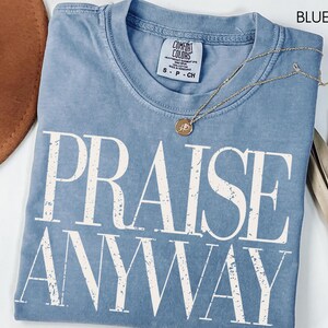 May include: A blue jean Comfort Colors t-shirt with the words "PRAISE ANYWAY" in large white letters. A gold necklace with a pendant rests on the shirt. A brown hat and dried plant are in the background.