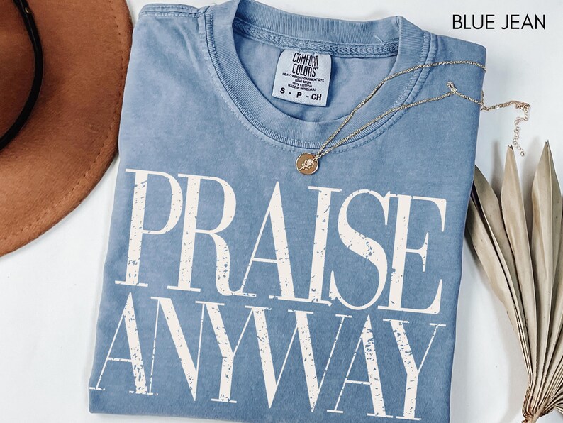 May include: A blue jean Comfort Colors t-shirt with the words "PRAISE ANYWAY" in large white letters. A gold necklace with a pendant rests on the shirt. A brown hat and dried plant are in the background.