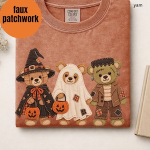 May include: A yam-colored t-shirt featuring a graphic of three teddy bears dressed for Halloween. One bear wears a witch hat and cape, one is a ghost, and one is a monster. Each bear holds a small orange jack-o'-lantern bucket.