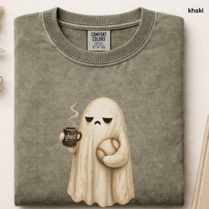 May include: A khaki-colored t-shirt featuring a graphic of a white ghost with a sad expression. The ghost holds a brown mug labeled ghost with steam rising and a baseball. The shirt has a Comfort Colors brand label inside the collar area.