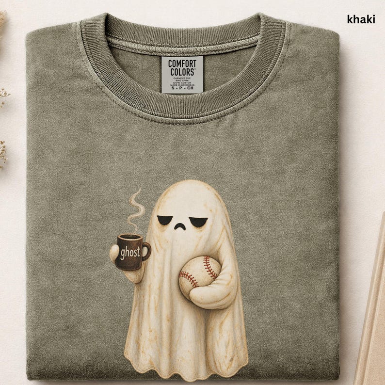 May include: A khaki-colored t-shirt featuring a graphic of a white ghost with a sad expression. The ghost holds a brown mug labeled ghost with steam rising and a baseball. The shirt has a Comfort Colors brand label inside the collar area.