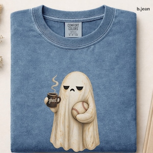 May include: A blue crew neck t-shirt with a Comfort Colors label at the neck. The graphic features a beige ghost with a sad expression holding a brown mug with the text ghost and a baseball. The ghost has steam rising from the mug.