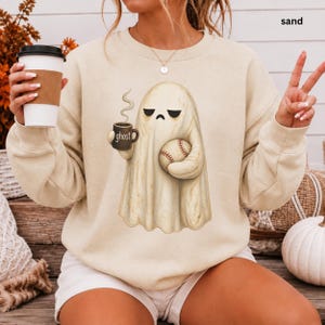May include: A sand-colored crewneck sweatshirt featuring a graphic of a ghost holding a steaming mug labeled ghost and a baseball. The ghost has a neutral expression with downward-curved eyes. The sweatshirt has long sleeves and a round neckline.