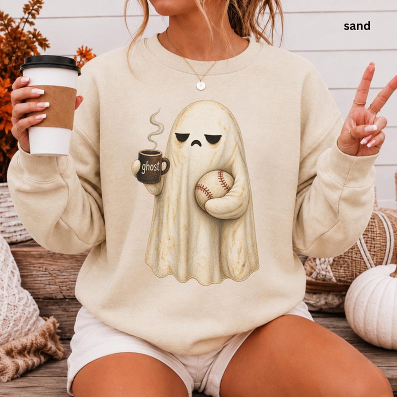 May include: A sand-colored crewneck sweatshirt featuring a graphic of a ghost holding a steaming mug labeled ghost and a baseball. The ghost has a neutral expression with downward-curved eyes. The sweatshirt has long sleeves and a round neckline.