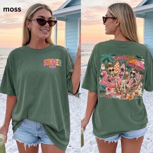May include: A moss green short-sleeve t-shirt featuring a small front graphic that reads "SUMMER WEEN" and a large back graphic depicting two skeletons on a beach with surfboards, a pink flamingo float, and the text "SUMMERWEEN" and "SPOOKY & SALTY".