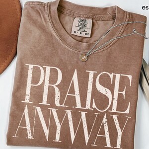 May include: Espresso-colored Comfort Colors t-shirt with the words "PRAISE ANYWAY" in a distressed, off-white font. A silver necklace with a small pendant rests on the shirt. The shirt is folded, and a brown hat and dried plant are visible.