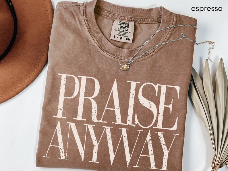 May include: Espresso-colored Comfort Colors t-shirt with the words "PRAISE ANYWAY" in a distressed, off-white font. A silver necklace with a small pendant rests on the shirt. The shirt is folded, and a brown hat and dried plant are visible.