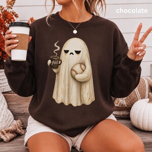 May include: A chocolate brown crewneck sweatshirt featuring a graphic of a cream-colored ghost with a sad expression. The ghost holds a dark mug labeled "ghost" with steam rising and a baseball. The text "chocolate" appears in the upper right corner.