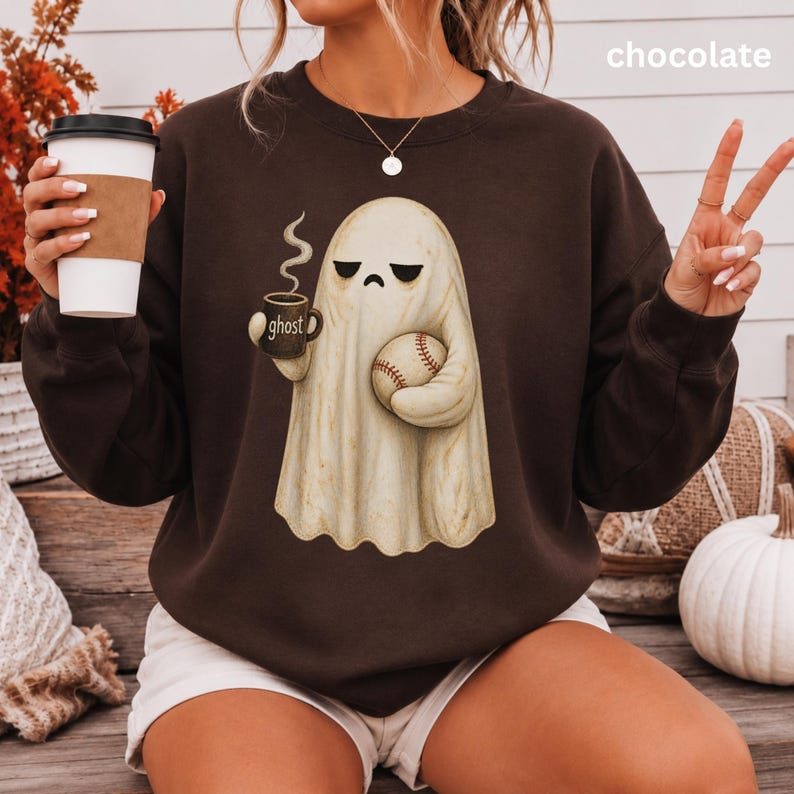 May include: A chocolate brown crewneck sweatshirt featuring a graphic of a cream-colored ghost with a sad expression. The ghost holds a dark mug labeled "ghost" with steam rising and a baseball. The text "chocolate" appears in the upper right corner.
