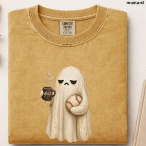 May include: A mustard yellow crew neck t-shirt with a Comfort Colors label. The graphic features a white ghost with a sad expression holding a brown mug labeled ghost with steam rising and a baseball in its other hand. The fabric has a textured look.