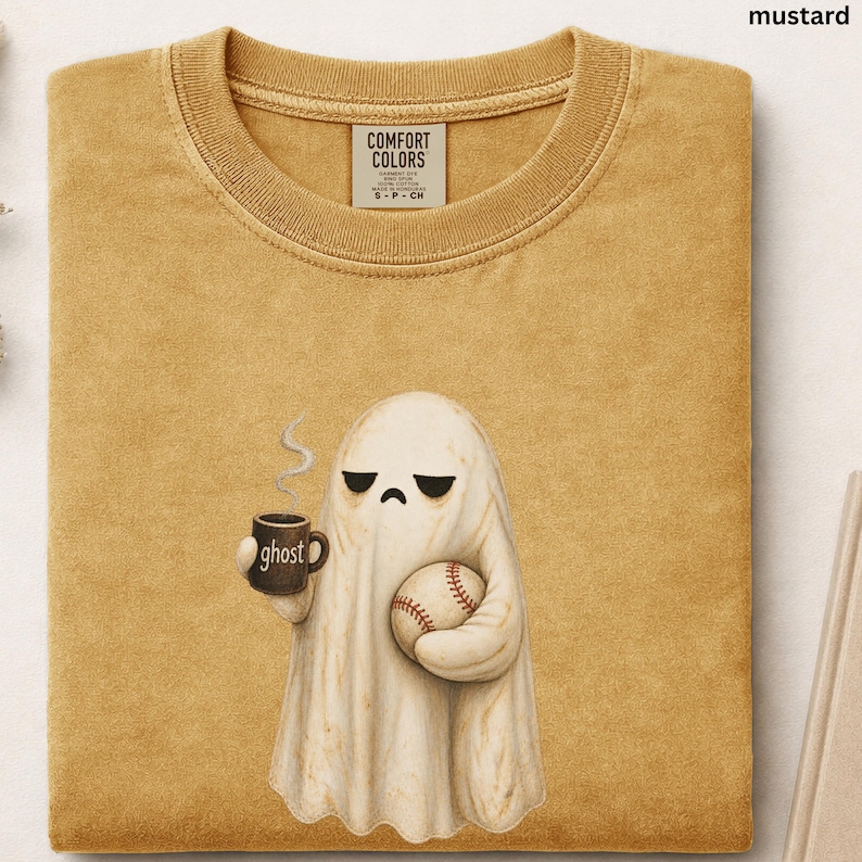 May include: A mustard yellow crew neck t-shirt with a Comfort Colors label. The graphic features a white ghost with a sad expression holding a brown mug labeled ghost with steam rising and a baseball in its other hand. The fabric has a textured look.