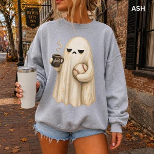 May include: An ash gray crewneck sweatshirt featuring a graphic of a cream-colored ghost with a grumpy expression. The ghost holds a black mug labeled ghost with steam rising and a baseball. The sweatshirt is paired with light blue denim shorts.