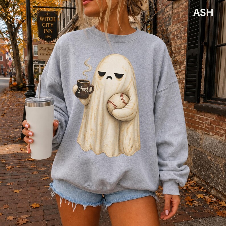 May include: An ash gray crewneck sweatshirt featuring a graphic of a cream-colored ghost with a grumpy expression. The ghost holds a black mug labeled ghost with steam rising and a baseball. The sweatshirt is paired with light blue denim shorts.