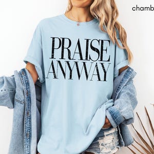 May include: Light blue t-shirt with the words "PRAISE ANYWAY" in a bold, black font. The shirt is part of a casual outfit, paired with a denim jacket. The word "chambray" is in the top right corner.
