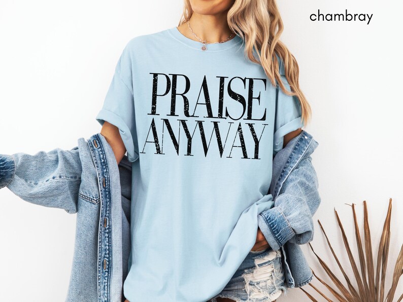 May include: Light blue t-shirt with the words "PRAISE ANYWAY" in a bold, black font. The shirt is part of a casual outfit, paired with a denim jacket. The word "chambray" is in the top right corner.