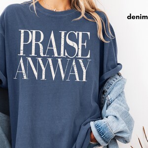 May include: A denim blue t-shirt with the words "PRAISE ANYWAY" in large, white, bold letters. The shirt has rolled-up sleeves and is paired with a denim jacket. The word "denim" is printed in the upper right corner.