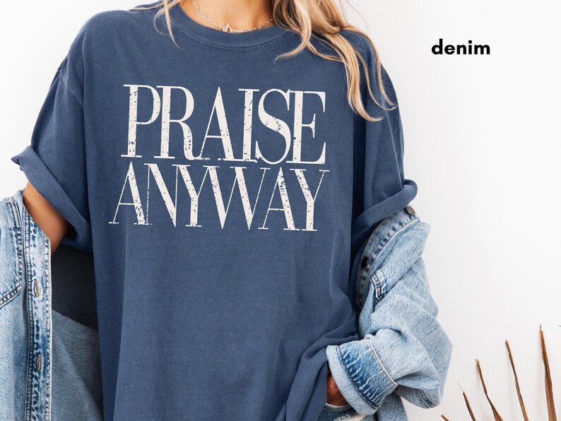 May include: A denim blue t-shirt with the words "PRAISE ANYWAY" in large, white, bold letters. The shirt has rolled-up sleeves and is paired with a denim jacket. The word "denim" is printed in the upper right corner.