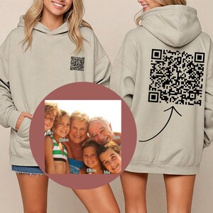 May include: A light gray hoodie with a black and white QR code on the back. The hoodie has a pocket on the front and a hood. A circle with a family photo is in the front of the hoodie. The photo shows a family of six people, including two young girls, two older women, and two older men. The names of the family members are written below their faces. The names are "Ian", "Chloe", "Olivia", "Mia", "Rob", and an unreadable name.