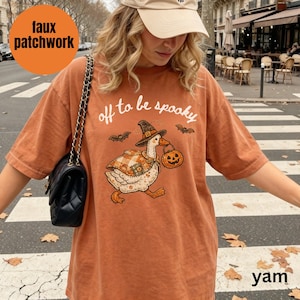 May include: A rust-colored short-sleeve t-shirt featuring a graphic of a goose wearing a witch hat and a plaid cape, carrying a jack-o'-lantern. The text "off to be spooky" is printed above the goose, with small bat illustrations on each side.