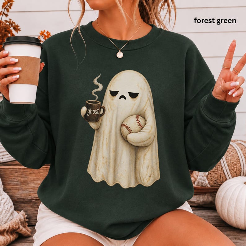 May include: A forest green crewneck sweatshirt featuring a graphic of a sad-looking white ghost holding a steaming mug labeled ghost and a baseball. The sweatshirt has long sleeves and a ribbed neckline, showcasing a casual and comfortable design.