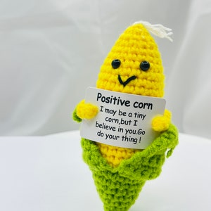 Könnte beinhalten: Eine gehäkelte Maispuppe, die ein Schild mit der Aufschrift "Positive corn I may be a tiny corn, but I believe in you. Go do your thing!" hält.