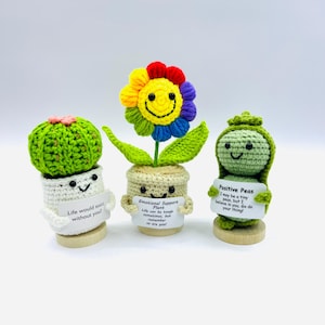 May include: Three crocheted plant figurines with smiling faces and motivational messages. A green cactus with the message "Life would suck without you!" A yellow flower with the message "Emotional Support Plant Life can be tough sometimes, but remember so are you!" and a green pea pod with the message "Positive Peas I may be a tiny peas, but I believe in you. Go do your thing!"