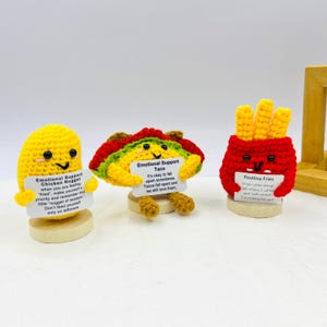 May include: Three crocheted food-themed plushies holding signs with encouraging messages. A yellow chicken nugget says "Emotional Support Chicken Nugget", a red and green taco says "Emotional Support Taco", and a red french fry container says "Positive Fries".