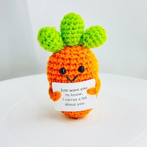 May include: A crocheted carrot toy with a green top and a smiling face. The carrot is holding a card that says "Just want you to know, I carrot a lot about you."
