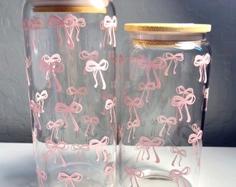 May include: Two glass jars with bamboo lids, both decorated with a pink bow pattern. The jars are clear and the bows are a light pink color.