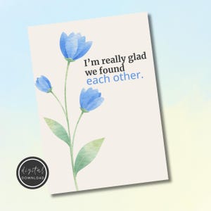 Può includere: Un biglietto di auguri con un design ad acquerello con fiori blu e il testo "I'm really glad we found each other". La carta ha uno sfondo beige e un logo di download digitale in basso a sinistra.