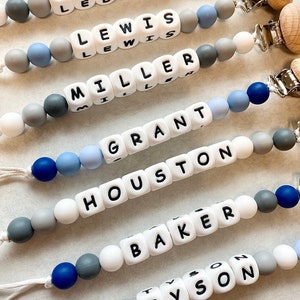 May include: Six silicone baby teethers with different names spelled out in black letters on white beads. The teethers are strung on a white cord with gray and blue beads. Each teether has a wooden clip attached.