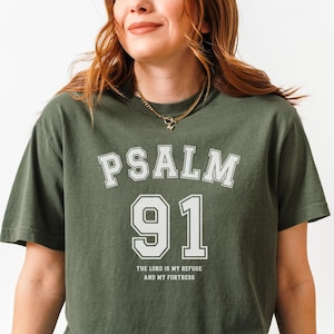 Può includere: T-shirt verde oliva con la scritta "PSALM 91" in lettere bianche e il numero "91" in un riquadro bianco. Sotto c'è la scritta "THE LORD IS MY REFUGE AND MY FORTRESS".