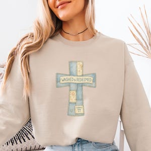 May include: A beige sweatshirt with a cross design. The cross is made of patchwork fabric in shades of blue and yellow, with the words "Washed & Redeemed" printed across it. The sweatshirt also has the Bible verse "1 Corinthians 6:11" printed on it.