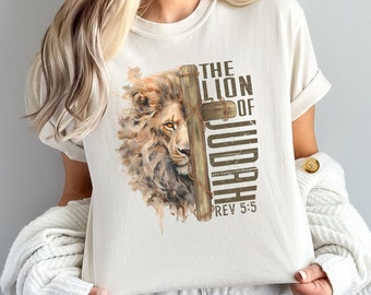 May include: Cream-colored t-shirt featuring a detailed illustration of a lion's face partially obscured by a wooden cross. The text "THE LION OF JUDAH REV 5:5" is printed vertically on the right side of the cross. The shirt has rolled-up sleeves.