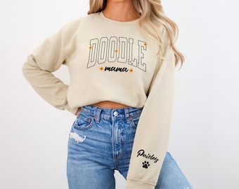 Puede incluir: Una sudadera beige recortada con el texto "Doodle Mama" en letras negras delineadas con pequeños acentos de flores amarillas. La manga izquierda tiene el texto "Paisley" en negro con una huella de pata.