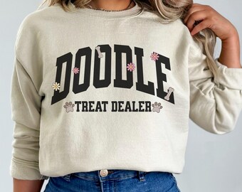 Puede incluir: Una sudadera beige con el texto "Doodle Treat Dealer" en negro con un hueso y huellas de patas. La palabra "Doodle" está en una fuente grande y en negrita con flores rosas y blancas.